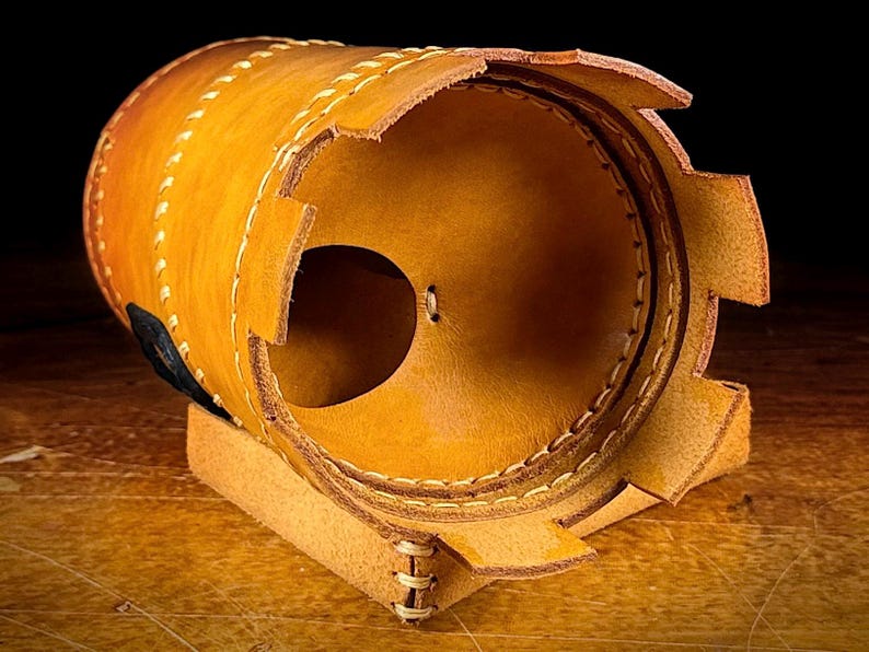 May include: A cylindrical leather object in tan and brown hues, featuring visible stitching and a circular opening. The leather has a textured appearance and is hand-stitched with a lighter thread. It rests on a small, rectangular base.