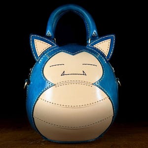 Pode incluir: Uma bolsa de couro azul e creme em forma de personagem de desenho animado. A bolsa apresenta um corpo arredondado, uma alça superior e detalhes costurados. O rosto do personagem é retratado na frente, com os olhos fechados e uma boca pequena.