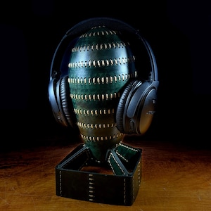 May include: A handcrafted leather headphone stand in shades of green and brown, stitched with gold thread. Black headphones rest on the stand, which is set on a wooden surface. The stand has a matching leather base.