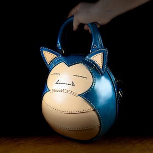 May include: A blue and tan leather handbag shaped like a cartoon character. The bag has a round body, two small handles, and features stitched details. The character's face is visible on the front, with closed eyes and a smiling mouth.