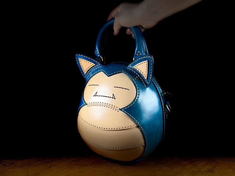 May include: A blue and tan leather handbag shaped like a cartoon character. The bag has a round body, two small handles, and features stitched details. The character's face is visible on the front, with closed eyes and a smiling mouth.