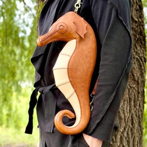 May include: A brown and tan seahorse-shaped crossbody bag with a long strap. The bag is made of leather and has detailed stitching. The seahorse design includes a curved tail and a detailed head. The bag is worn over the shoulder.
