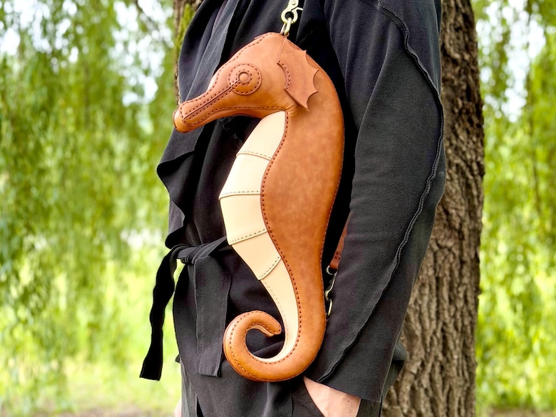 May include: A brown and tan seahorse-shaped crossbody bag with a long strap. The bag is made of leather and has detailed stitching. The seahorse design includes a curved tail and a detailed head. The bag is worn over the shoulder.