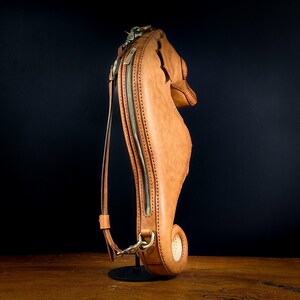 May include: A tan leather seahorse-shaped bag with a long strap and a brass zipper. The bag has a detailed design, including stitching and a curved tail. The bag is standing upright on a black stand against a dark background.