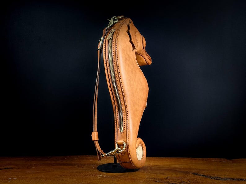 May include: A tan leather seahorse-shaped bag with a long strap and a brass zipper. The bag has a detailed design, including stitching and a curved tail. The bag is standing upright on a black stand against a dark background.