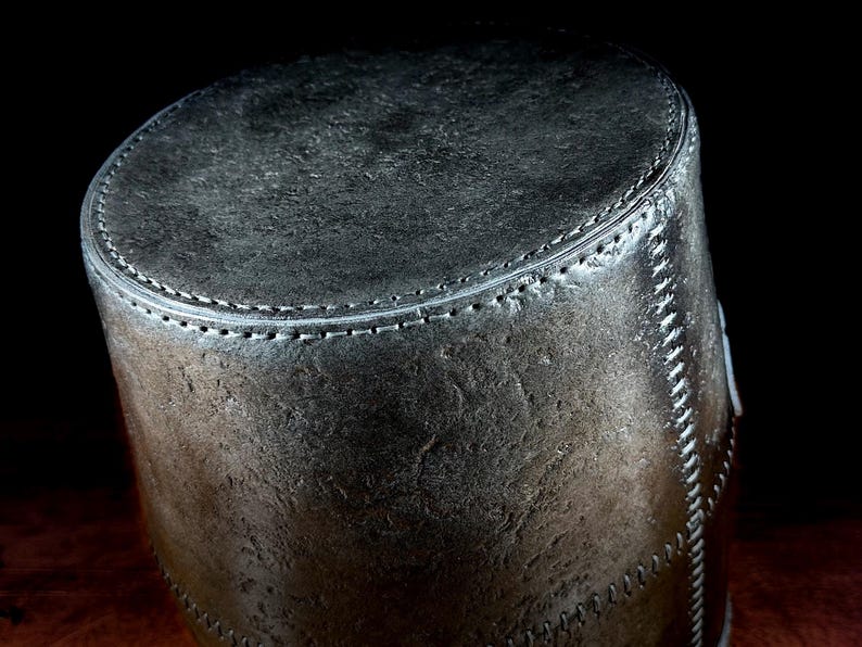 K&ouml;nnte beinhalten: Ein zylindrischer, dunkelgrauer Lederbeh&auml;lter mit strukturierter Oberfl&auml;che. Die oberen und seitlichen N&auml;hte sind mit wei&szlig;em Faden gen&auml;ht. Der Beh&auml;lter hat ein robustes, Vintage-Aussehen.