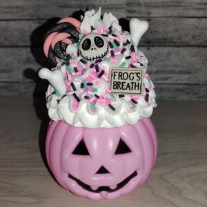 Puede incluir: Contenedor en forma de calabaza rosa con glaseado blanco, chispas rosas y verdes, un bast&oacute;n de caramelo blanco y negro, una cara de Jack Skellington en blanco y negro y un letrero que dice "FROG'S BREATH".