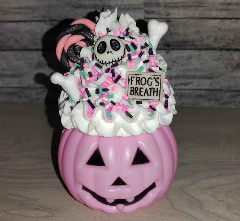 Puede incluir: Contenedor en forma de calabaza rosa con glaseado blanco, chispas rosas y verdes, un bast&oacute;n de caramelo blanco y negro, una cara de Jack Skellington en blanco y negro y un letrero que dice "FROG'S BREATH".