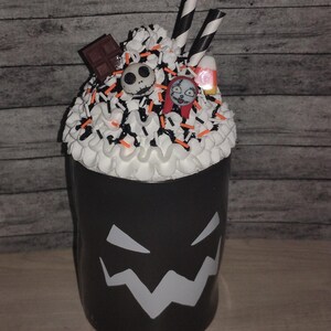 May include: A black cup with a white, jagged face design. The cup is topped with white frosting, sprinkles, and candy. Decorations include a chocolate bar, Jack Skellington and Sally figures, and candy corn. Two black and white striped straws are in the frosting.