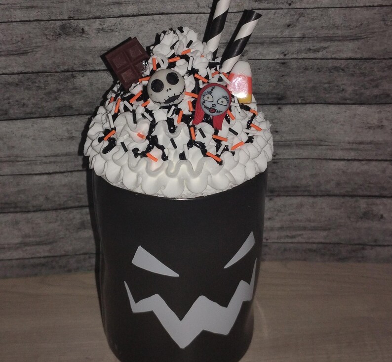 May include: A black cup with a white, jagged face design. The cup is topped with white frosting, sprinkles, and candy. Decorations include a chocolate bar, Jack Skellington and Sally figures, and candy corn. Two black and white striped straws are in the frosting.