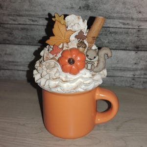 May include: A ceramic mug with a white whipped cream topping decorated with autumn-themed accents, including a small squirrel, a pumpkin, and leaves. The mug is orange and has a handle.