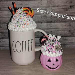 Puede incluir: Comparaci&oacute;n de tama&ntilde;o de dos tazas de caf&eacute; con tem&aacute;tica de Halloween. La taza m&aacute;s grande es blanca con la palabra "COFFEE" impresa en ella. Est&aacute; cubierta con crema batida blanca, chispas y un bast&oacute;n de caramelo. La taza m&aacute;s peque&ntilde;a es una calabaza rosa con una cara de Jack Skellington y un letrero de "FROGS BREATH". Tambi&eacute;n est&aacute; cubierta con crema batida, chispas y un bast&oacute;n de caramelo.