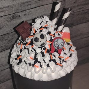 May include: A Halloween-themed dessert with white frosting, black and orange sprinkles, and decorative elements. The dessert features a Jack Skellington face, Sally face, chocolate square, candy corn, and black and white striped straws.