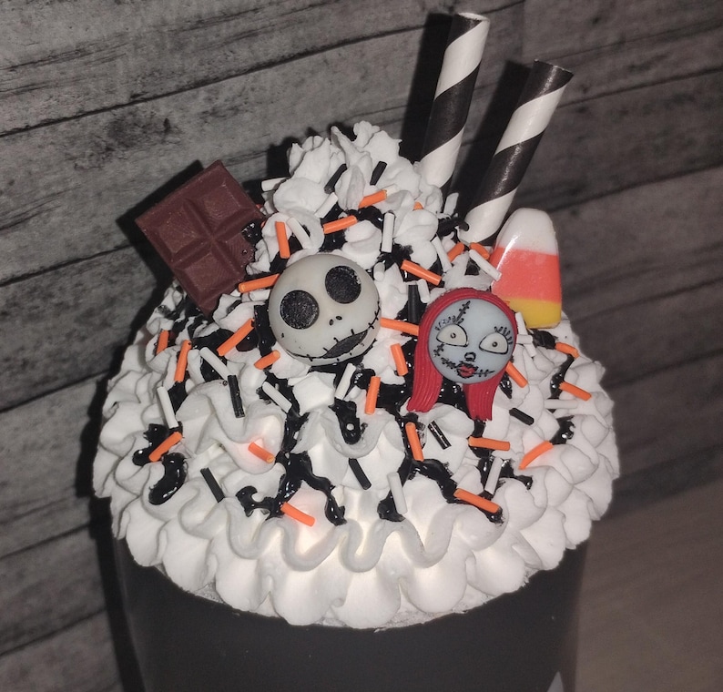 May include: A Halloween-themed dessert with white frosting, black and orange sprinkles, and decorative elements. The dessert features a Jack Skellington face, Sally face, chocolate square, candy corn, and black and white striped straws.