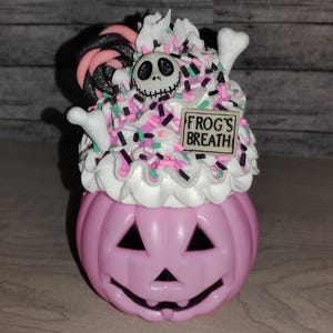 Puede incluir: Recipiente de color rosa en forma de calabaza con una cobertura blanca similar a la cobertura de glaseado. La cobertura est&aacute; decorada con chispas negras y rosas, un bast&oacute;n de caramelo verde y blanco y un peque&ntilde;o hueso blanco. Un peque&ntilde;o letrero blanco con el texto "FROG'S BREATH" est&aacute; en la parte superior de la cobertura. Se puede ver una cara de Jack Skellington en blanco y negro en la cobertura.