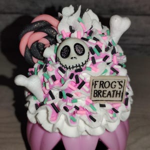 Puede incluir: Un recipiente en forma de calabaza rosa con glaseado blanco y espolvoreado. El glaseado est&aacute; cubierto con una cara de Jack Skellington en blanco y negro y una piruleta de espiral rosa y negro. El recipiente tiene un letrero que dice "FROG'S BREATH".