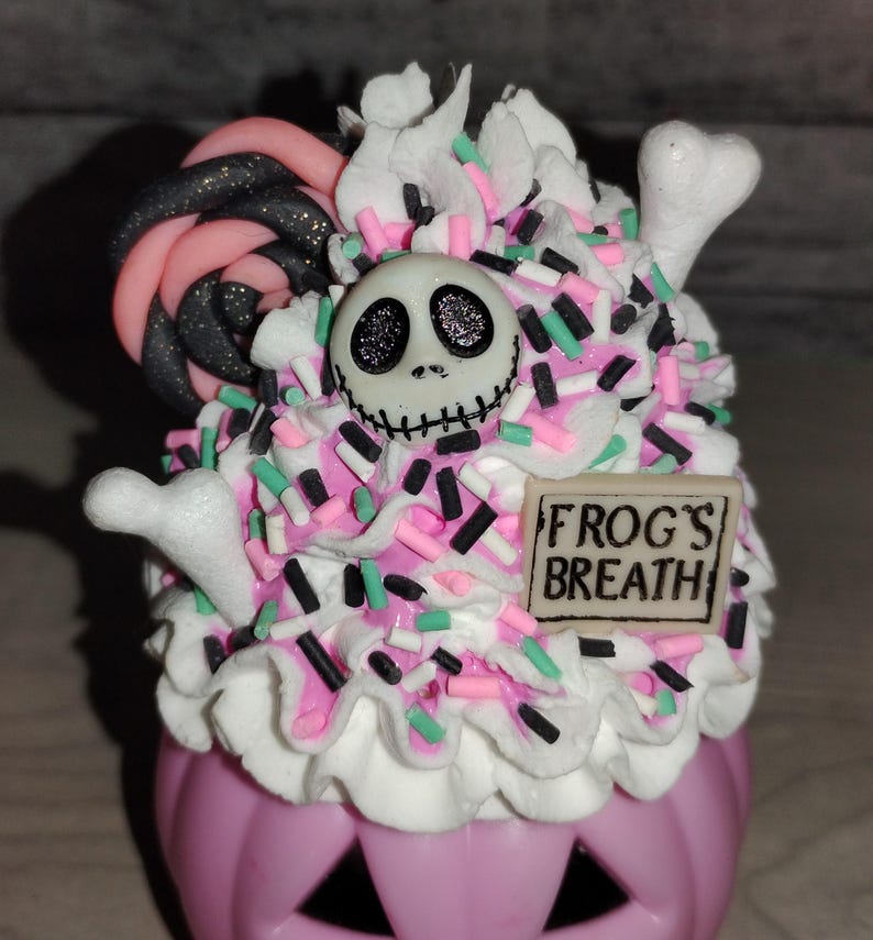 Puede incluir: Un recipiente en forma de calabaza rosa con glaseado blanco y espolvoreado. El glaseado est&aacute; cubierto con una cara de Jack Skellington en blanco y negro y una piruleta de espiral rosa y negro. El recipiente tiene un letrero que dice "FROG'S BREATH".