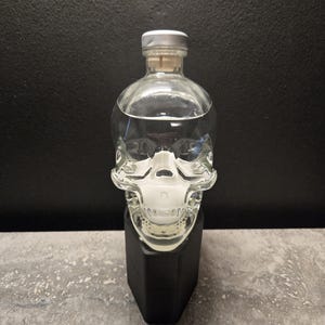 May include: A clear glass skull-shaped bottle with a silver cap and cork stopper. The bottle is filled with a clear liquid and sits on a black, geometric base. The background is a dark gray.