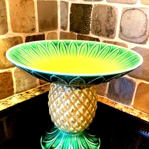 May include: A green and yellow ceramic pineapple-shaped bowl with a wide, shallow bowl on top. The bowl is decorated with a green and yellow pineapple design.