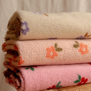 Puede incluir: Una pila de tres mantas de forro polar sherpa dobladas con una superficie texturizada y difusa. La manta superior es crema con flores moradas, la del medio es melocotón claro con flores naranjas y la inferior es rosa claro con flores rojas.