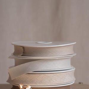 May include: A stack of four circular spools of fabric ribbon in varying shades of neutral beige, tan, and brown. Each spool features a woven texture and a flat edge, with the ribbon ends neatly wound around white plastic cores in a vertical column.