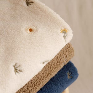 May include: A stack of three soft blankets in cream, brown, and navy blue. The cream blanket has embroidered yellow and brown flowers. The brown and navy blankets are solid colours. The blankets are folded and stacked.