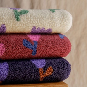 May include: Three rolled fleece blankets stacked on top of each other. The top blanket is cream-colored with green and purple floral patterns. The middle blanket is burgundy with pink and blue floral patterns. The bottom blanket is dark purple with orange and purple floral patterns.