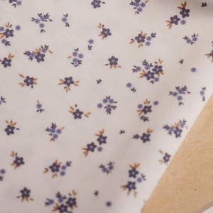 Op de afbeelding: Witte stof met een delicaat bloemmotief met kleine blauwe bloemen en bruine bladeren. De stof is gedrapeerd over een lichtgekleurd houten oppervlak, dat de herhaling van het patroon laat zien.