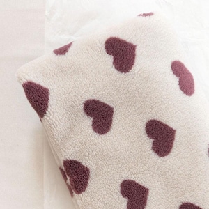 Puede incluir: Una manta plegada de color crema con una textura de felpa. La manta está decorada con formas de corazón de color burdeos oscuro espaciadas uniformemente. La manta está doblada cuidadosamente, mostrando el patrón de corazón en su superficie.