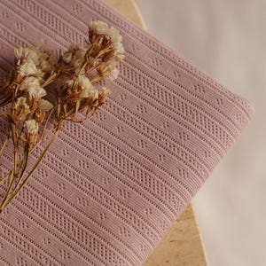 May include: Close-up of a folded, dusty rose fabric with a detailed, geometric pattern. The fabric is adorned with a sprig of dried, cream-colored flowers. The texture of the fabric is visible, highlighting its intricate design.