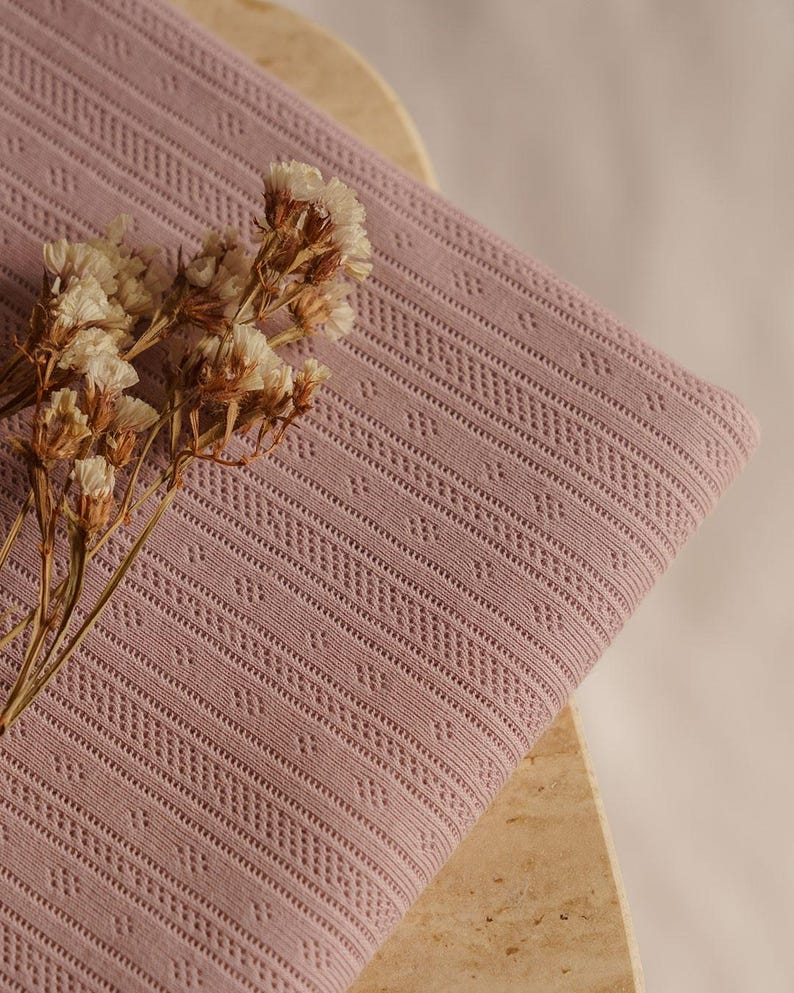 May include: Close-up of a folded, dusty rose fabric with a detailed, geometric pattern. The fabric is adorned with a sprig of dried, cream-colored flowers. The texture of the fabric is visible, highlighting its intricate design.