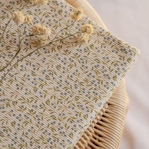 May include: A close-up shot of folded fabric with a floral design. The fabric features a cream backdrop with a repeating pattern of small blue dots and green leaves. Dried flowers are placed on top of the fabric, which is resting on a wicker surface.