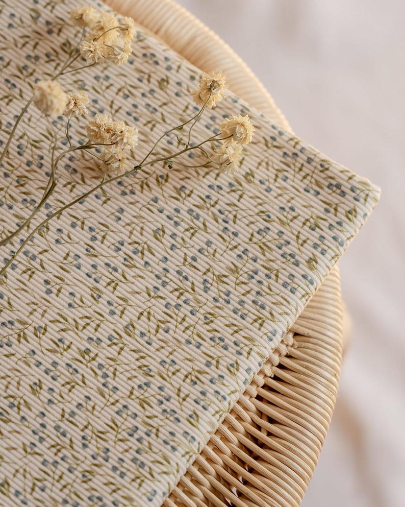 May include: A close-up shot of folded fabric with a floral design. The fabric features a cream backdrop with a repeating pattern of small blue dots and green leaves. Dried flowers are placed on top of the fabric, which is resting on a wicker surface.