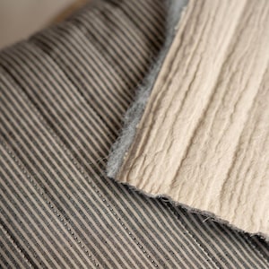May include: A textile piece featuring a dark gray and white pinstripe pattern on one side and a solid beige, textured, crinkled fabric on the other. The beige section has a visible, fuzzy, light gray edge where the two distinct fabrics meet.
