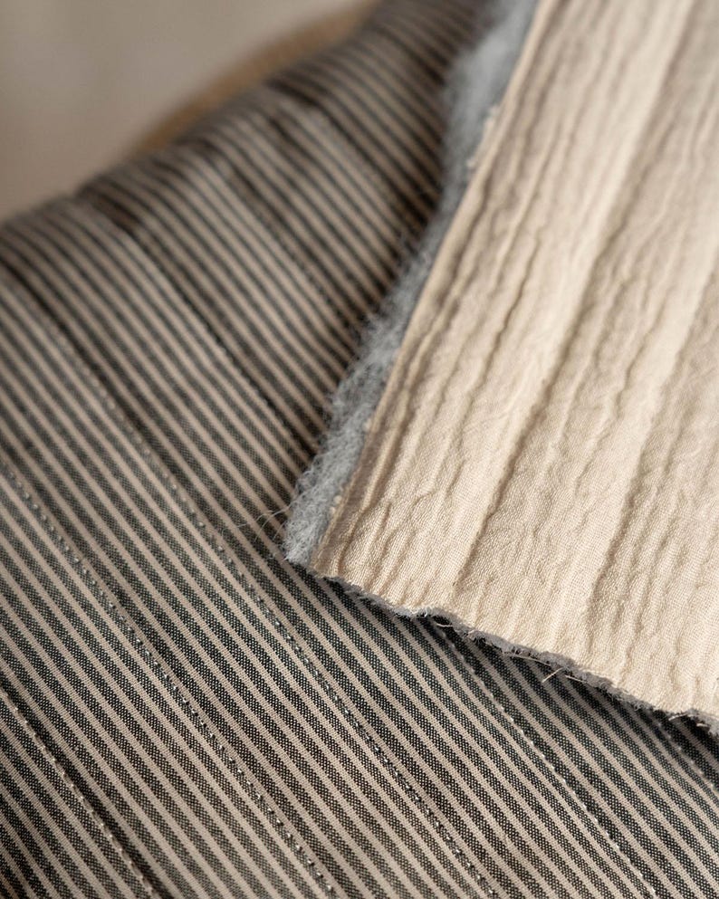 May include: A textile piece featuring a dark gray and white pinstripe pattern on one side and a solid beige, textured, crinkled fabric on the other. The beige section has a visible, fuzzy, light gray edge where the two distinct fabrics meet.