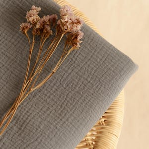 Op de afbeelding: Een gevouwen, grijze stof met een getextureerd oppervlak, mogelijk mousseline of gaas, rust op een geweven rieten stoel. Gedroogde, lichtroze bloemen met bruine stelen zijn op de stof geplaatst. De achtergrond is lichtbeige.