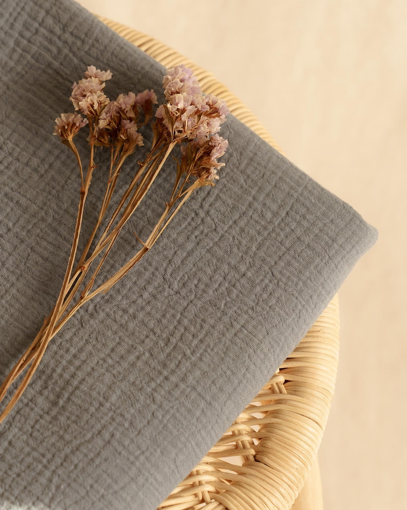 Op de afbeelding: Een gevouwen, grijze stof met een getextureerd oppervlak, mogelijk mousseline of gaas, rust op een geweven rieten stoel. Gedroogde, lichtroze bloemen met bruine stelen zijn op de stof geplaatst. De achtergrond is lichtbeige.