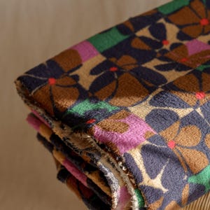 May include: Folded floral patterned blanket. The blanket features a repeating pattern of brown, navy, pink, and green flowers with red centers. The blanket is folded and appears to be made of a soft, textured material.
