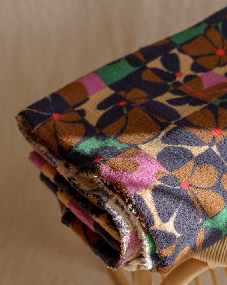 May include: Folded floral patterned blanket. The blanket features a repeating pattern of brown, navy, pink, and green flowers with red centers. The blanket is folded and appears to be made of a soft, textured material.