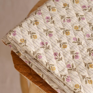 May include: A folded, quilted blanket with a floral pattern of yellow and pink flowers on a cream background. The blanket has a striped design with thin purple lines. The edge of the blanket is a solid brown color.