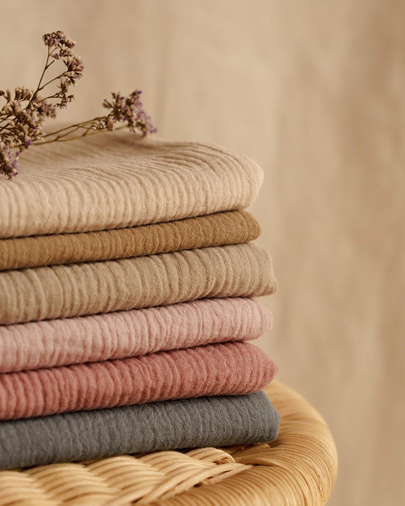 Op de afbeelding: Een stapel van zes gevouwen, getextureerde stoffen in verschillende neutrale kleuren: beige, bruin, lichtbruin, roze, rozerood en grijs. Een takje gedroogde paarse bloemen ligt erbovenop. De stoffen zijn gestapeld op een geweven, lichtbruin oppervlak.
