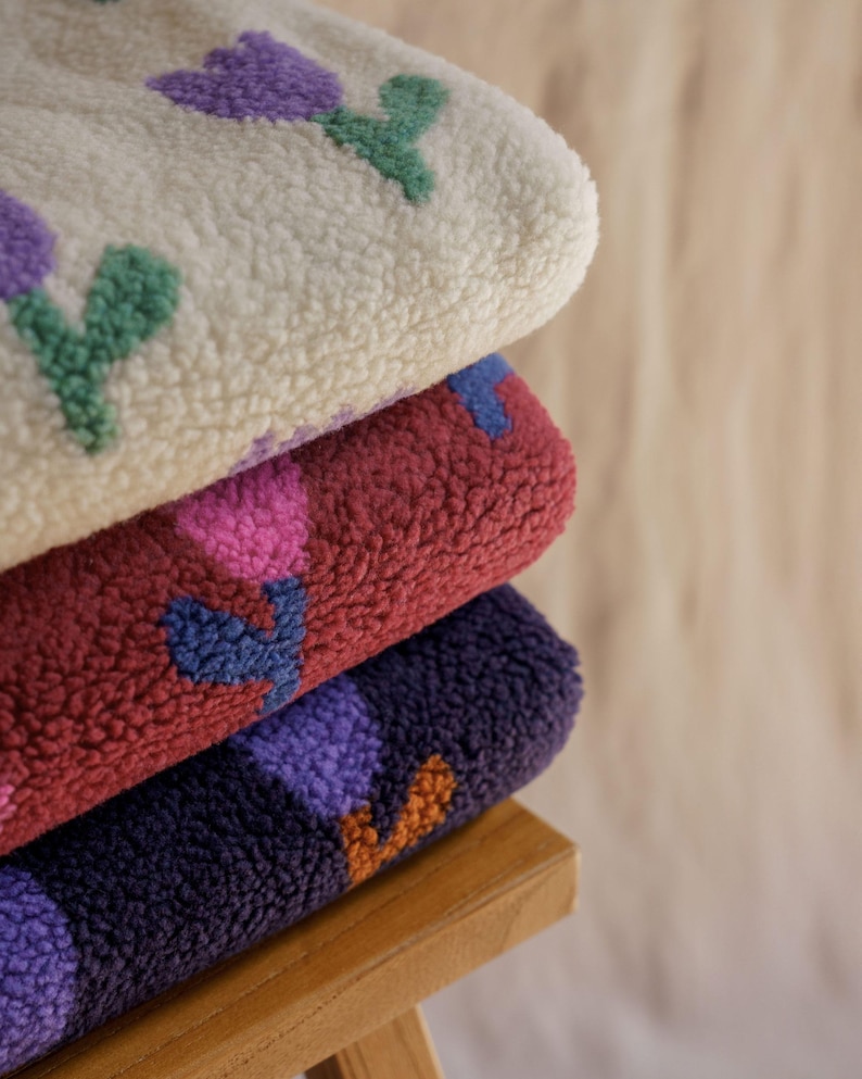 May include: A stack of three plush, textured blankets. The top blanket is cream-colored with purple and green floral designs. The middle blanket is burgundy with pink and blue flowers. The bottom blanket is dark purple with orange and purple flowers.