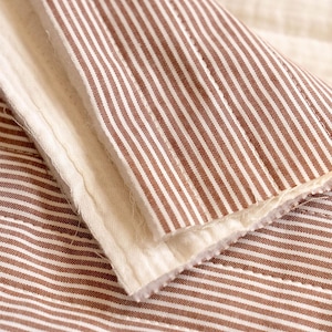 May include: A folded textile featuring a pattern of thin, vertical brown and white stripes. The fabric has a quilted texture with visible stitching lines and a soft, off-white interior lining visible along the folded edge of the material.