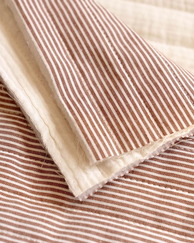 May include: A folded textile featuring a pattern of thin, vertical brown and white stripes. The fabric has a quilted texture with visible stitching lines and a soft, off-white interior lining visible along the folded edge of the material.