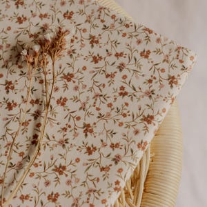 May include: A folded piece of fabric with a cream background and a delicate floral pattern in shades of brown and pink. The fabric is resting on a woven wicker surface, with dried flowers in the foreground.