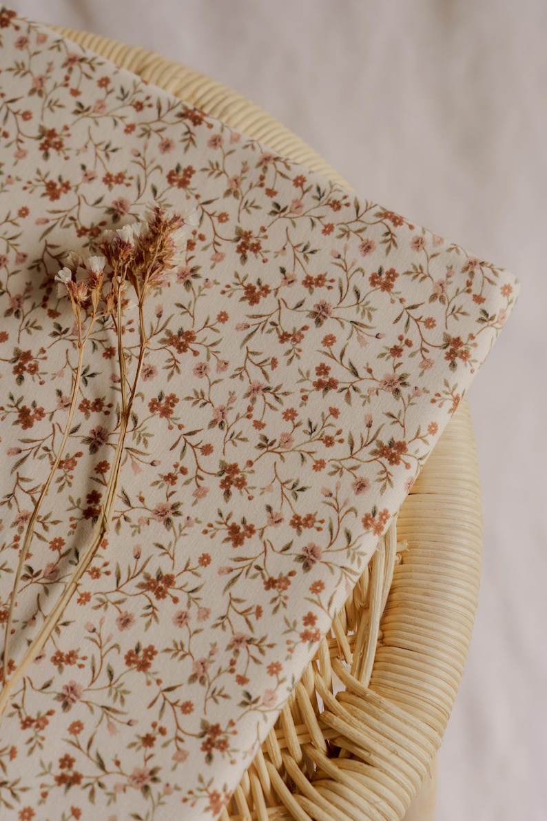 May include: A folded piece of fabric with a cream background and a delicate floral pattern in shades of brown and pink. The fabric is resting on a woven wicker surface, with dried flowers in the foreground.