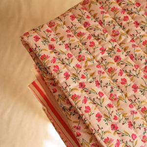 Op de afbeelding: Gevouwen, gewatteerde stof met een bloemenpatroon. De stof heeft een lichtroze achtergrond met een herhalend patroon van kleine rode bloemen en groene bladeren. De randen zijn afgewerkt met een gestreepte stof.