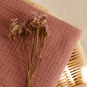Op de afbeelding: Een close-up van een stoffen textuur in de kleur oudroze, mogelijk mousseline of gaas, gedrapeerd over een rotan stoel. Gedroogde, lichtpaarse bloemen liggen op de stof, wat een zachte, natuurlijke uitstraling cre&euml;ert. De afbeelding heeft een warme, uitnodigende uitstraling.