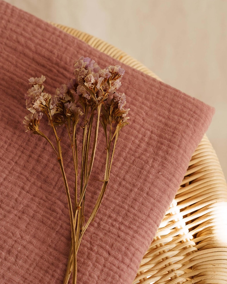 Op de afbeelding: Een close-up van een stoffen textuur in de kleur oudroze, mogelijk mousseline of gaas, gedrapeerd over een rotan stoel. Gedroogde, lichtpaarse bloemen liggen op de stof, wat een zachte, natuurlijke uitstraling cre&euml;ert. De afbeelding heeft een warme, uitnodigende uitstraling.