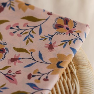 May include: A folded piece of light pink fabric featuring a repeating floral pattern. The design includes stylized flowers and leaves in shades of mustard yellow, dark blue, and muted pink, arranged across the soft-toned background surface.